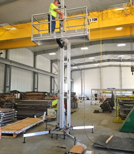 Worker operating ReechCraft PowerMast freestanding system inside an industrial workshop, standing on elevated platform supported by aluminum mast with stabilizing outriggers.