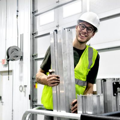 Person wearing safety vest and hard hat assembling aluminum mast sections of ReechCraft PowerLift inside a workshop with metal garage doors