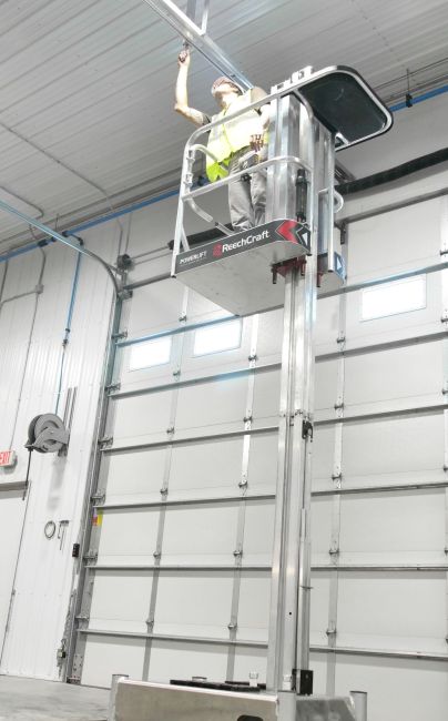 Worker on ReechCraft PowerLift PL-X performing overhead maintenance inside a large industrial garage with high ceilings and sectional doors.