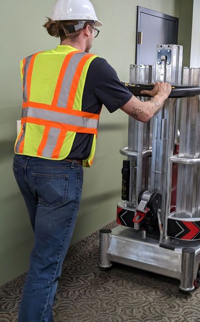 Worker in safety vest and hard hat maneuvering ReechCraft PowerLift PL-X Compact Base through an indoor hallway.