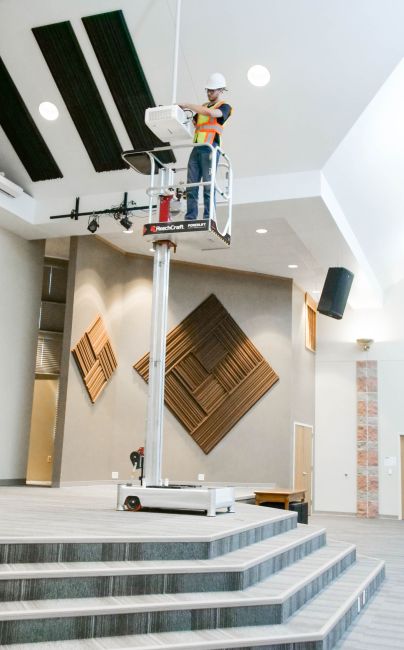 Worker using ReechCraft PowerLift PL-X Compact Base to perform overhead maintenance in a modern auditorium.