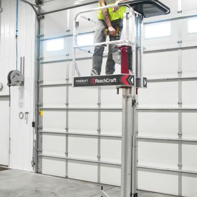 Person standing on ReechCraft PowerLift inside a garage or workshop, elevated platform with outriggers and wheels for stability during overhead work
