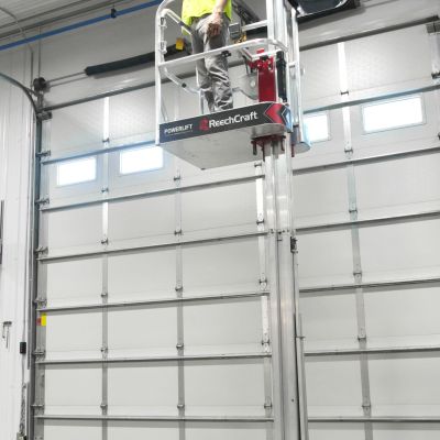 Worker standing on ReechCraft PowerLift inside a large garage performing overhead maintenance near ceiling beams and lighting fixtures
