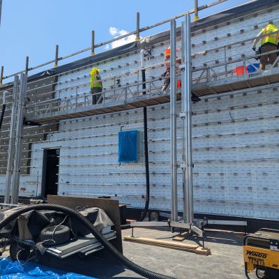 ReechCraft PowerMast freestanding system supporting a long work platform with multiple workers performing exterior wall installation on a commercial building under clear sky.