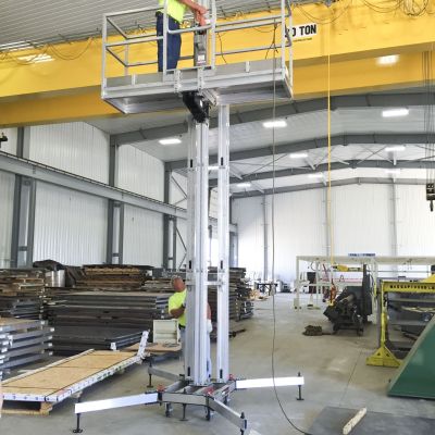 Worker standing on ReechCraft PowerMast freestanding system platform inside an industrial workshop, elevated for overhead maintenance with aluminum mast and stabilizing outriggers.