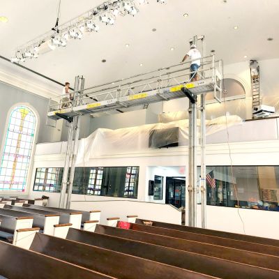 Two workers standing on extended ReechCraft PowerMast freestanding system platform inside a church sanctuary, positioned above pews for overhead lighting and ceiling maintenance.