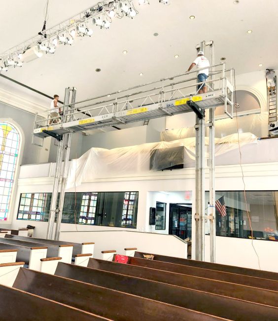 Two workers using ReechCraft PowerMast freestanding system with extended platform inside a large church sanctuary, positioned above pews for ceiling and lighting maintenance.