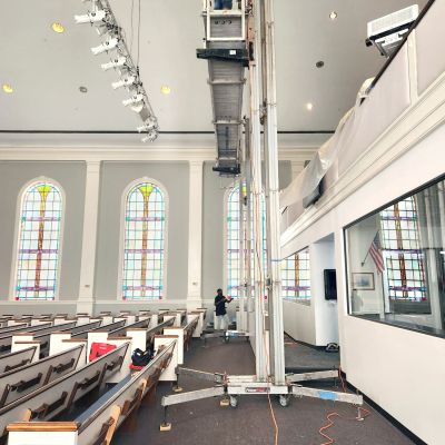 ReechCraft PowerMast freestanding system set up inside a church sanctuary, with extended platform reaching ceiling for lighting maintenance above pews and stained glass windows.
