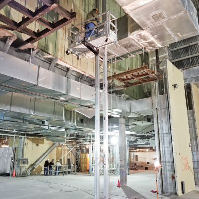 ReechCraft PowerMast freestanding system in an industrial building, with dual masts supporting a work platform elevated near large HVAC ducts for overhead installation and maintenance.