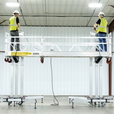 Two workers operating ReechCraft PowerMast freestanding system in a warehouse, standing on an extended aluminum platform supported by dual mast configurations with stabilizing outriggers.