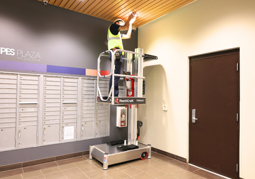 A worker is elevated on a PowerLift platform near the ceiling, performing maintenance or installation work.