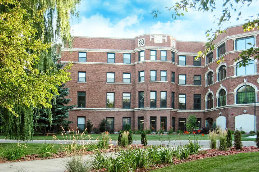 Red brick multi-story building with large rectangular windows and decorative architectural details, set around a landscaped courtyard with grass, ornamental plants, and a curved walkway, partially framed by leafy trees under a bright blue sky.