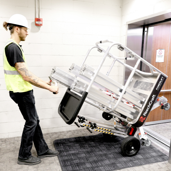 Worker wearing a hard hat and safety vest controlling a tilted ReechCraft PowerLift on wheels near an elevator, demonstrating compact transport and maneuverability indoors.