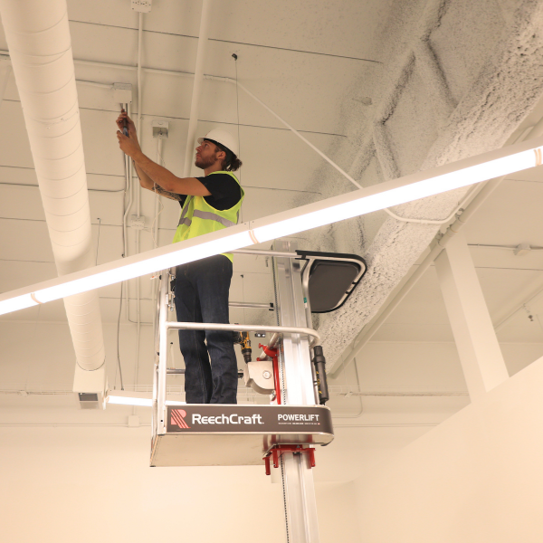 Worker in a hard hat and high-visibility vest using a ReechCraft PowerLift to perform overhead work on a ceiling inside a building.
