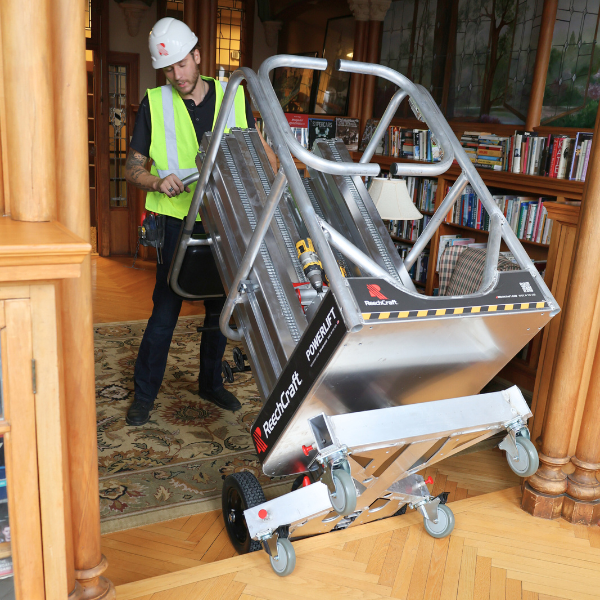 Worker wearing a hard hat and safety vest carefully steering a ReechCraft PowerLift mobile lift over a threshold into a finished indoor space with wood floors, bookshelves, and tools on the platform.