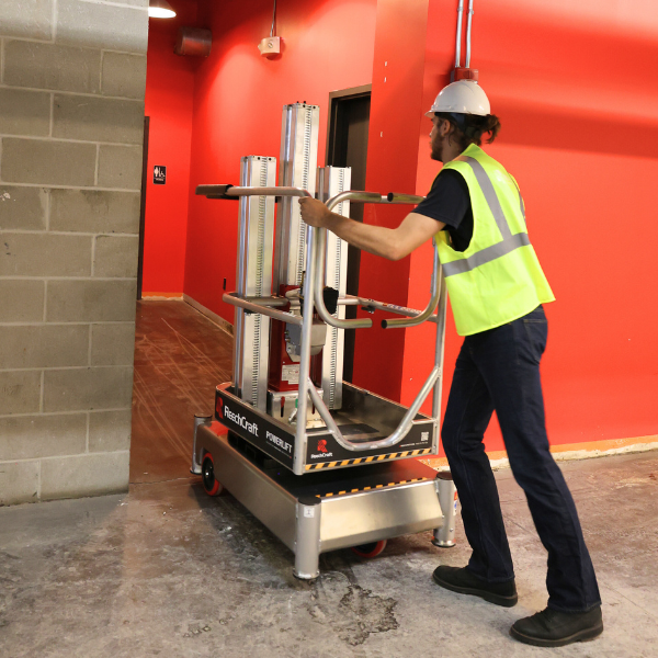 Worker wearing a hard hat and safety vest pushing a ReechCraft PowerLift lift system in an indoor construction area.